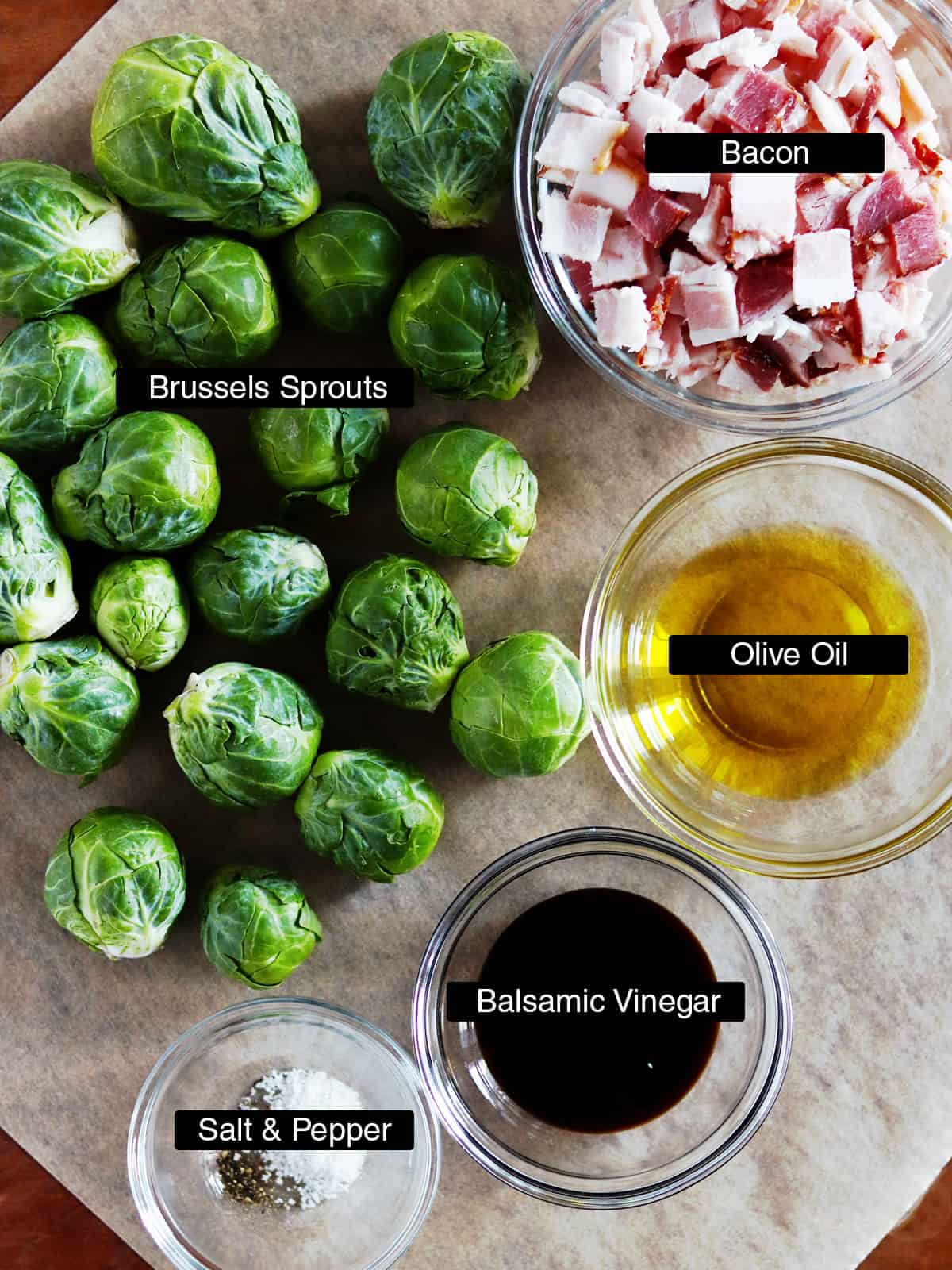 Ingredients for roasted Brussels sprouts and bacon on a parchment paper lined brown table.