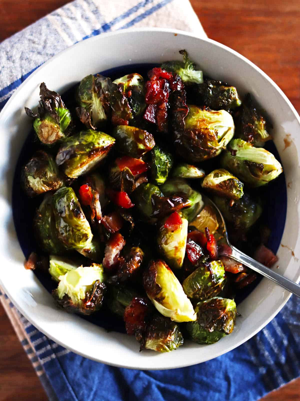 Roasted Brussels sprouts and bacon in a ceramic bowl with a blue and tan plaid napkin.