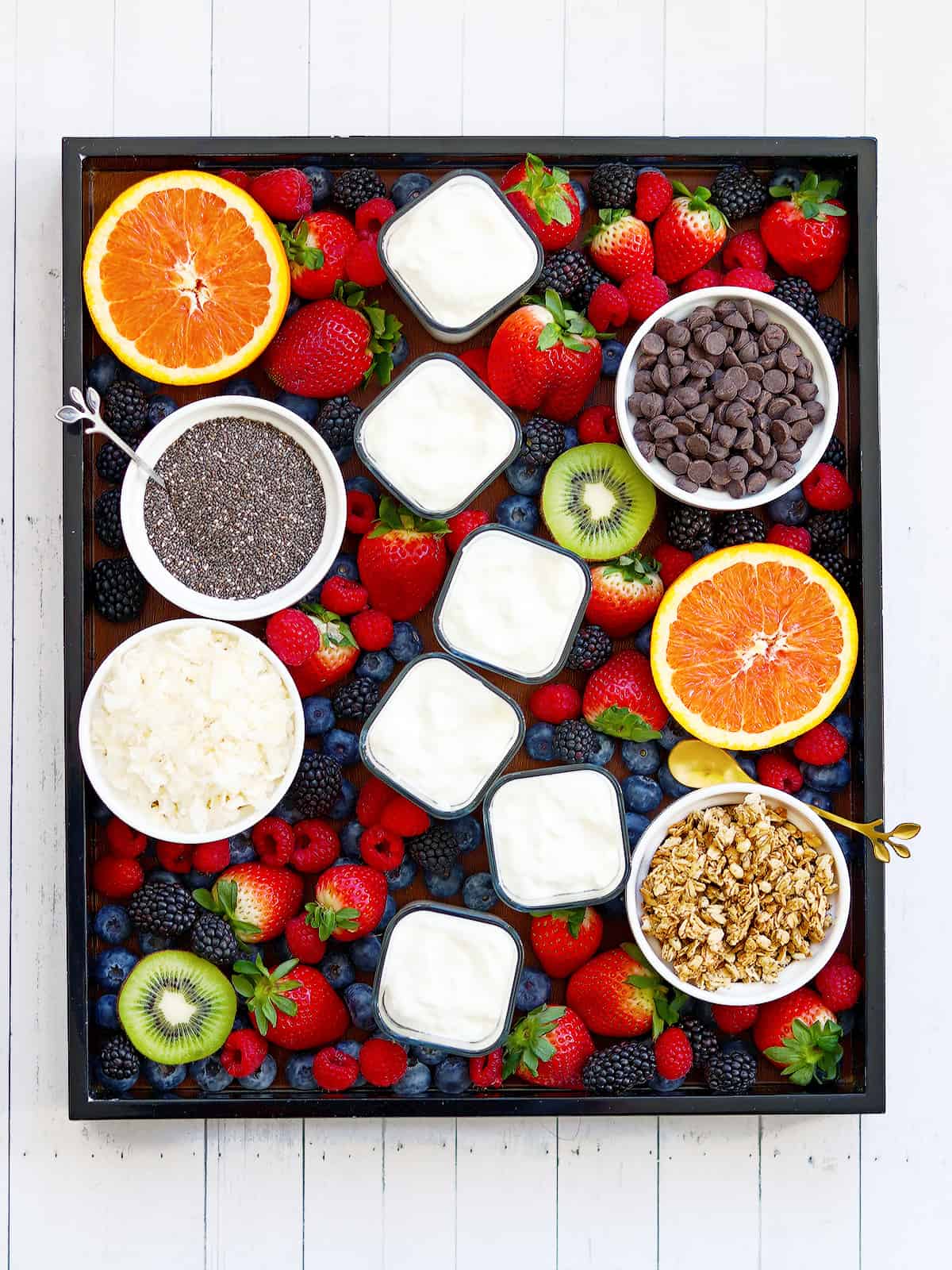 Yogurt charcuterie board with individual yogurt cups, fruit and toppings on a white wood table.