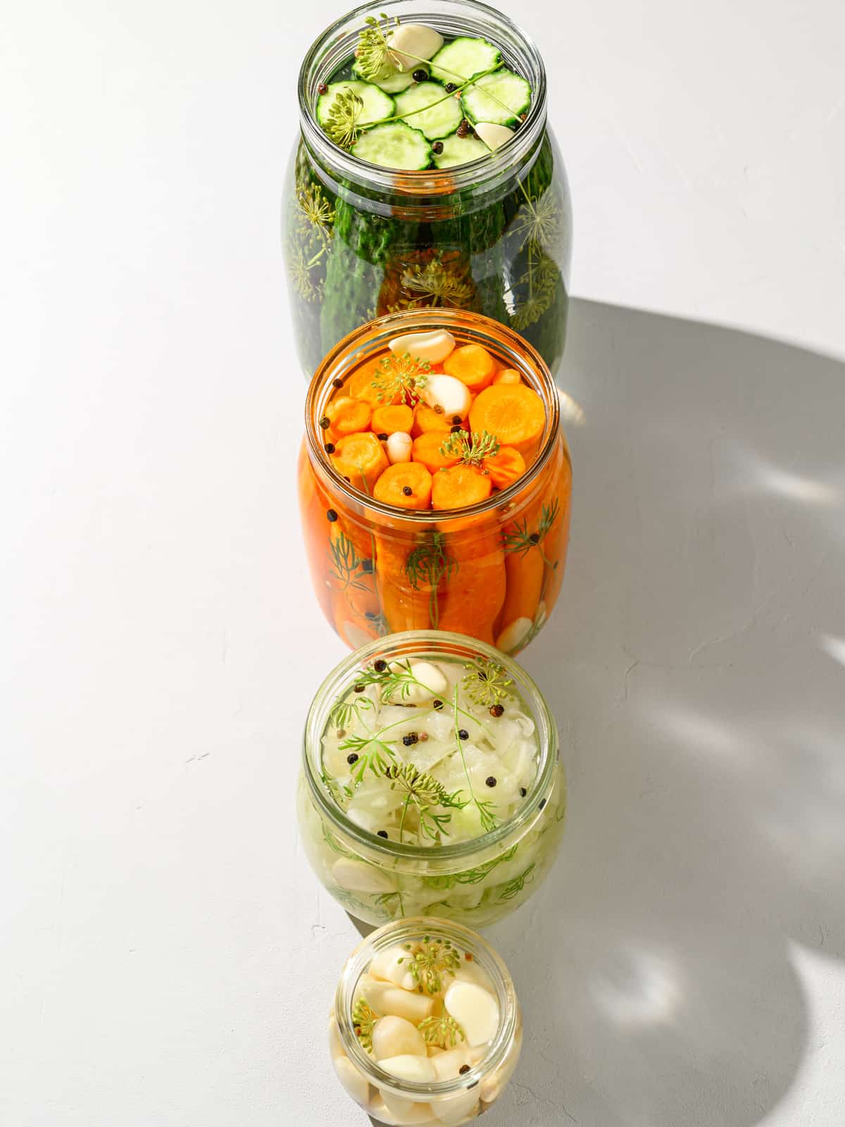 Homemade pickles, fermented carrots, sauerkraut and fermented garlic in glass jars on a white tabletop.