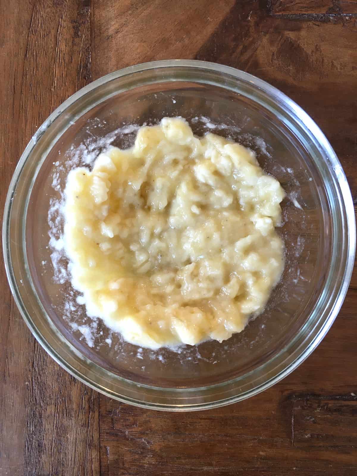 Mashed bananas in a glass mixing bowl on a brown wood table.