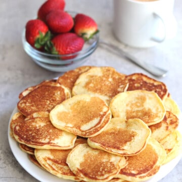 Mini pancakes on a white plate with a bowl of fresh strawberries.