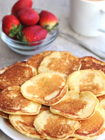 Mini pancakes on a white plate with a bowl of fresh strawberries.