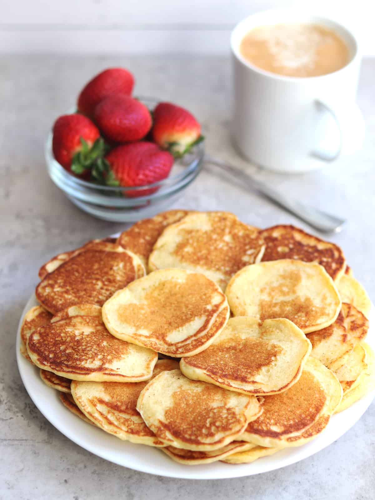 Mini pancakes on a white plate with a bowl of fresh strawberries and a cup of espresso in the background.