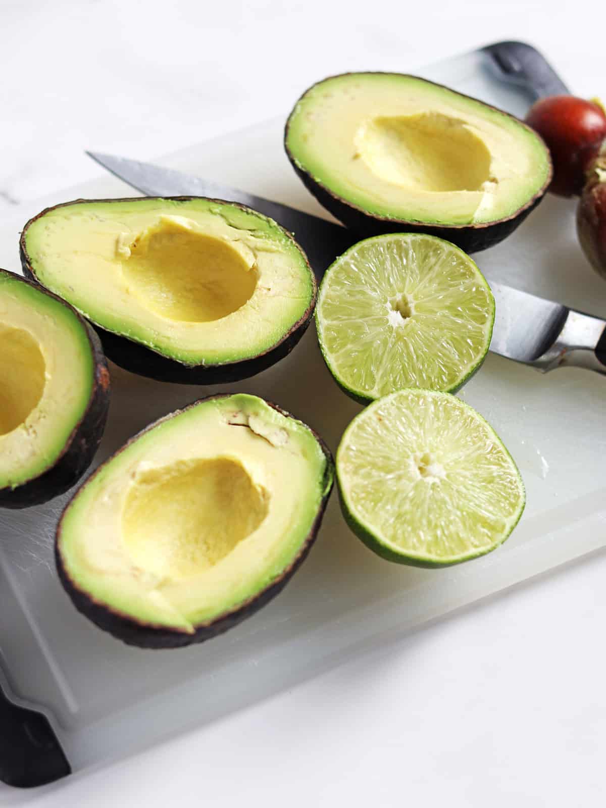 Halved avocados and a halved lime with a sharp knife on a white and black cutting board.