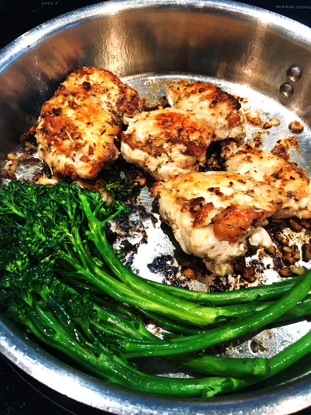 Pan seared chicken thighs and broccolini in a stainless steel skillet on a black glass stove top.