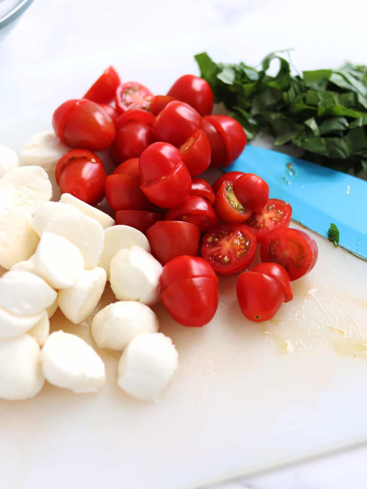Halved baby mozzarella balls, cherry tomatoes and sliced basil on a white cutting board with a light blue ceramic knife.