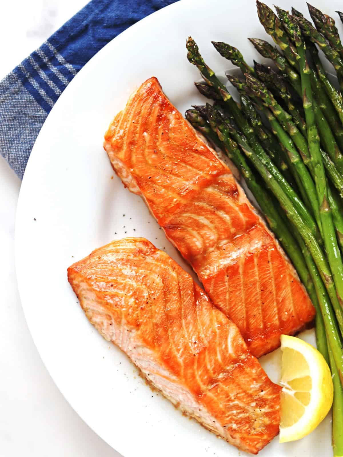 Oven baked salmon fillets on a white plate with a side of asparagus and a slice of lemon.