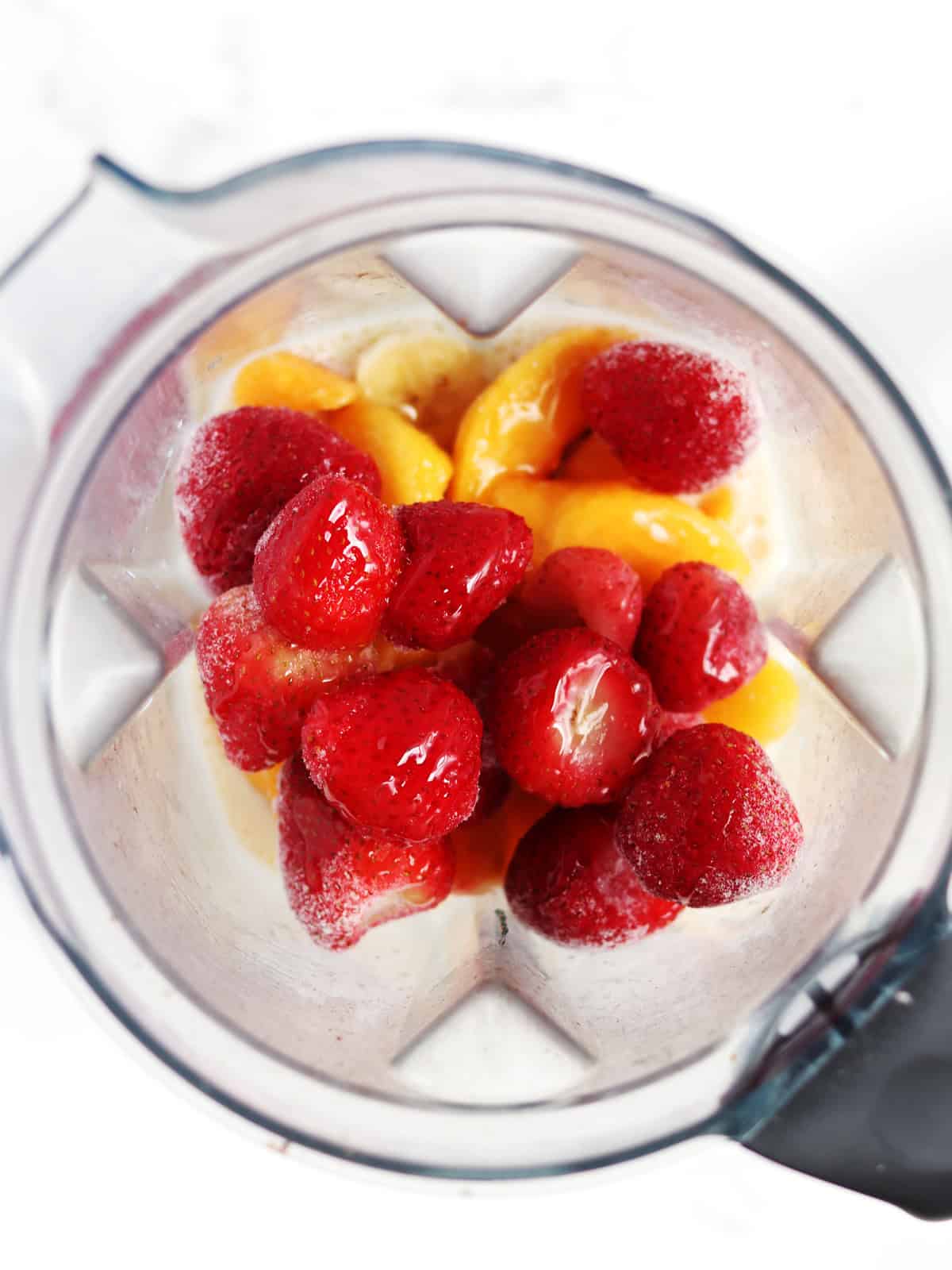 Frozen strawberries, peaches, honey, banana and soy milk in a blender on a white marble countertop.