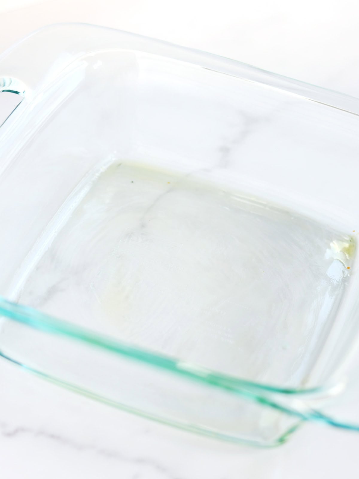 Glass baking dish greased with a thin layer of olive oil atop a white marble countertop.