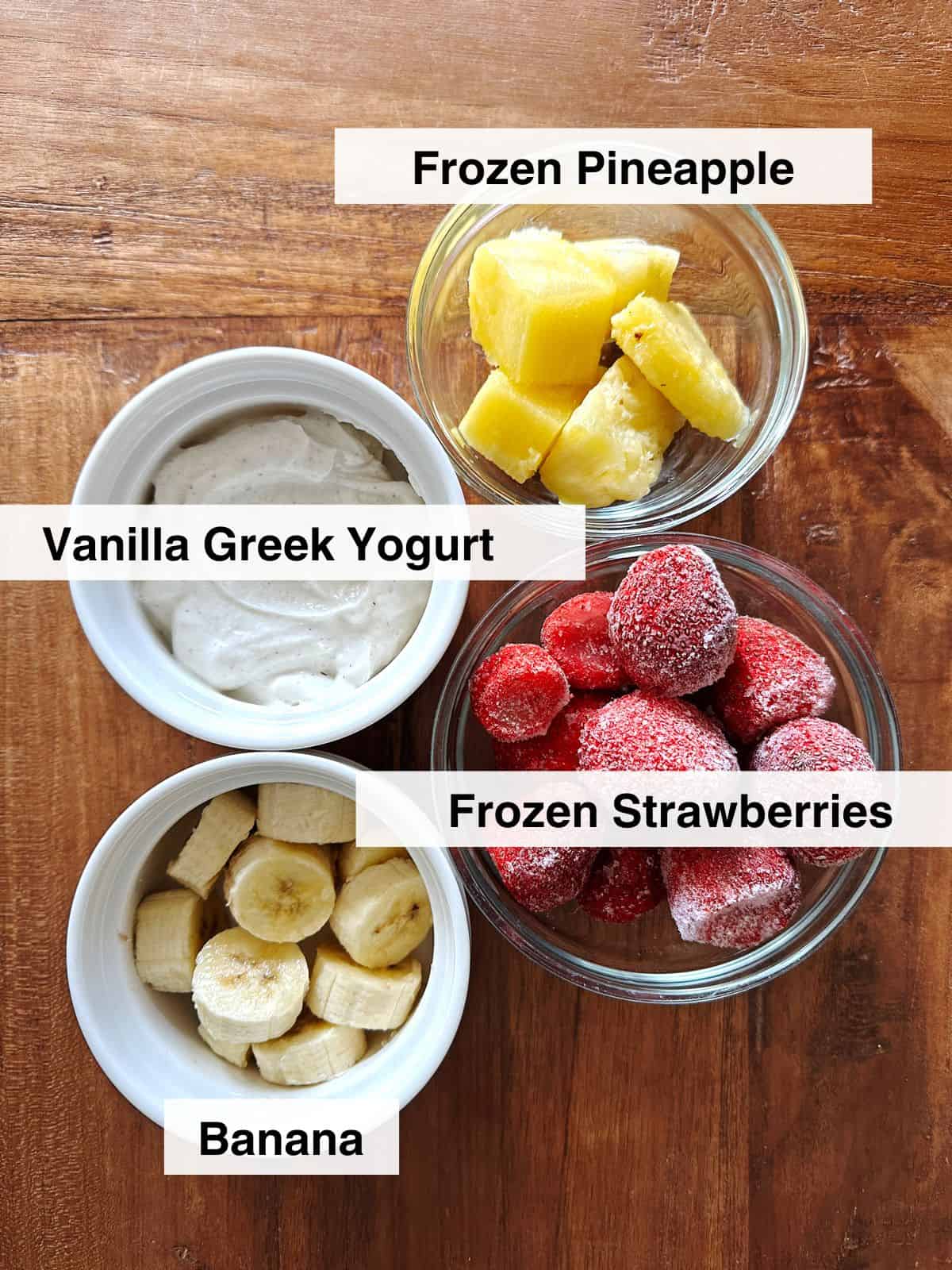 Vanilla Greek yogurt, chopped banana, frozen pineapple and frozen strawberries in meal prep dishes on a brown wood table.