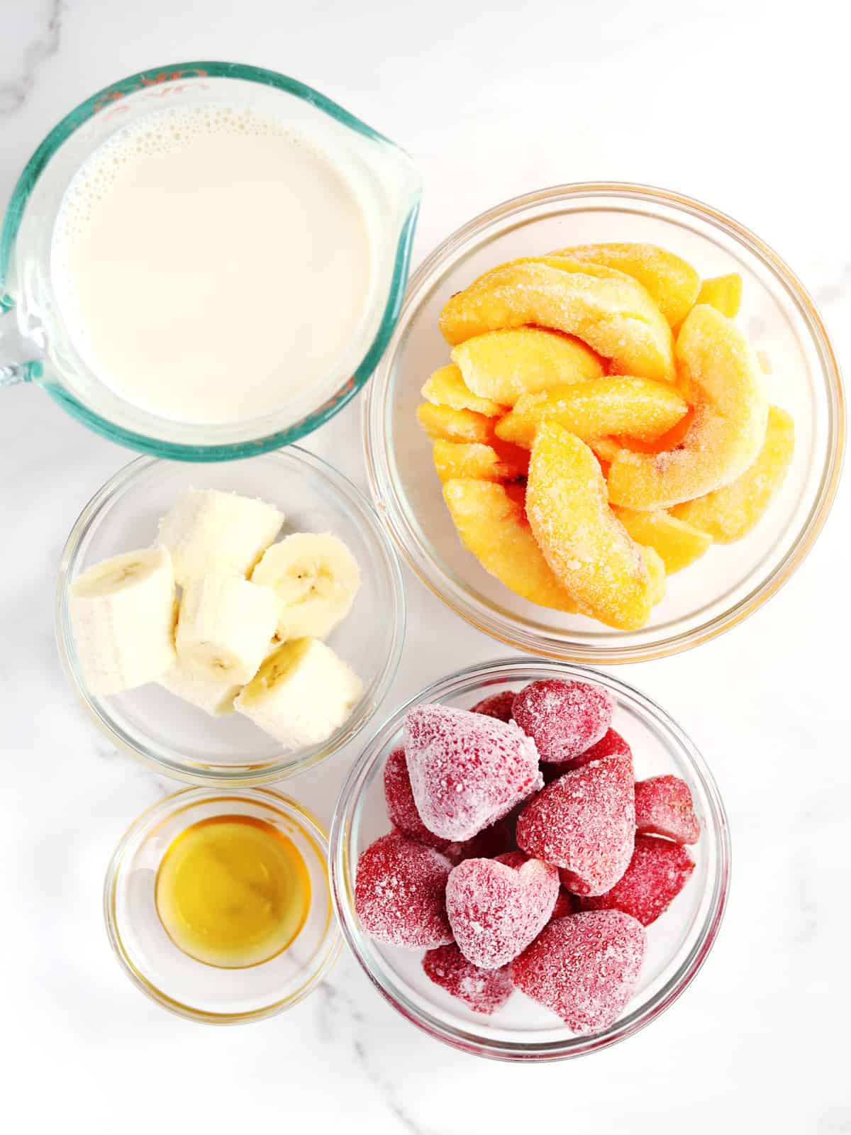 Strawberry peach smoothie ingredients in glass meal prep bowls on a white marble countertop.