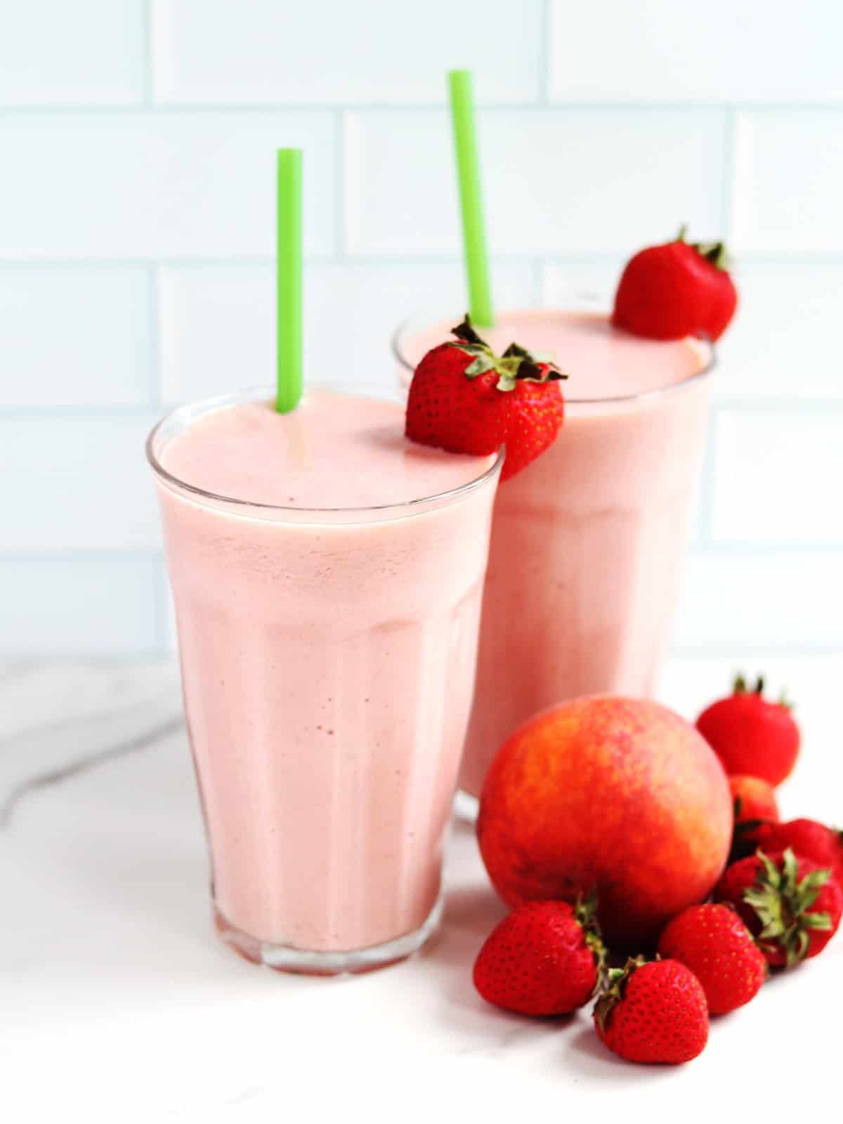 Two large strawberry peach smoothies with green straws on a white marble countertop.