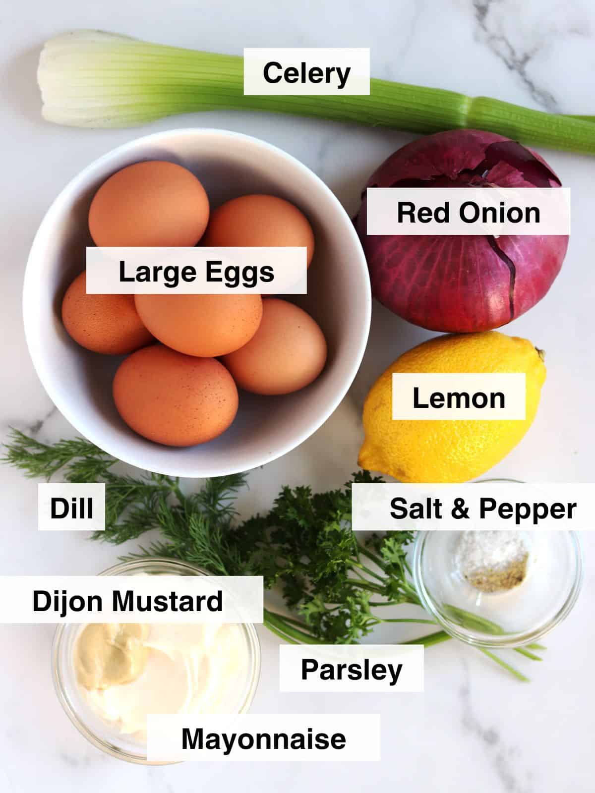 A bowl of large eggs surrounded by labeled ingredients: celery, red onion, lemon, dill, salt & pepper, Dijon mustard, parsley, and mayonnaise, all arranged on a white surface.