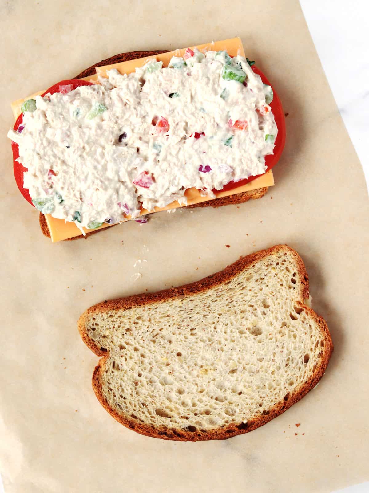 A slice of bread topped with American cheese, tomato slices, and a creamy tuna salad with a second bread slice next to it, ready to close the sandwich.