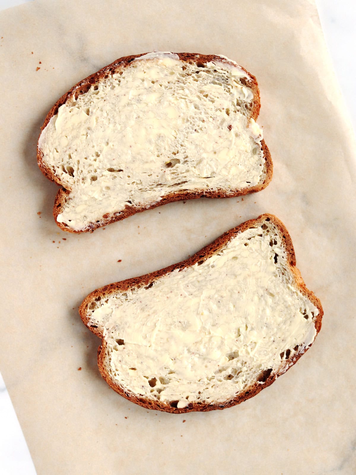 Two slices of whole grain bread with butter spread evenly on top, placed on a sheet of parchment paper.
