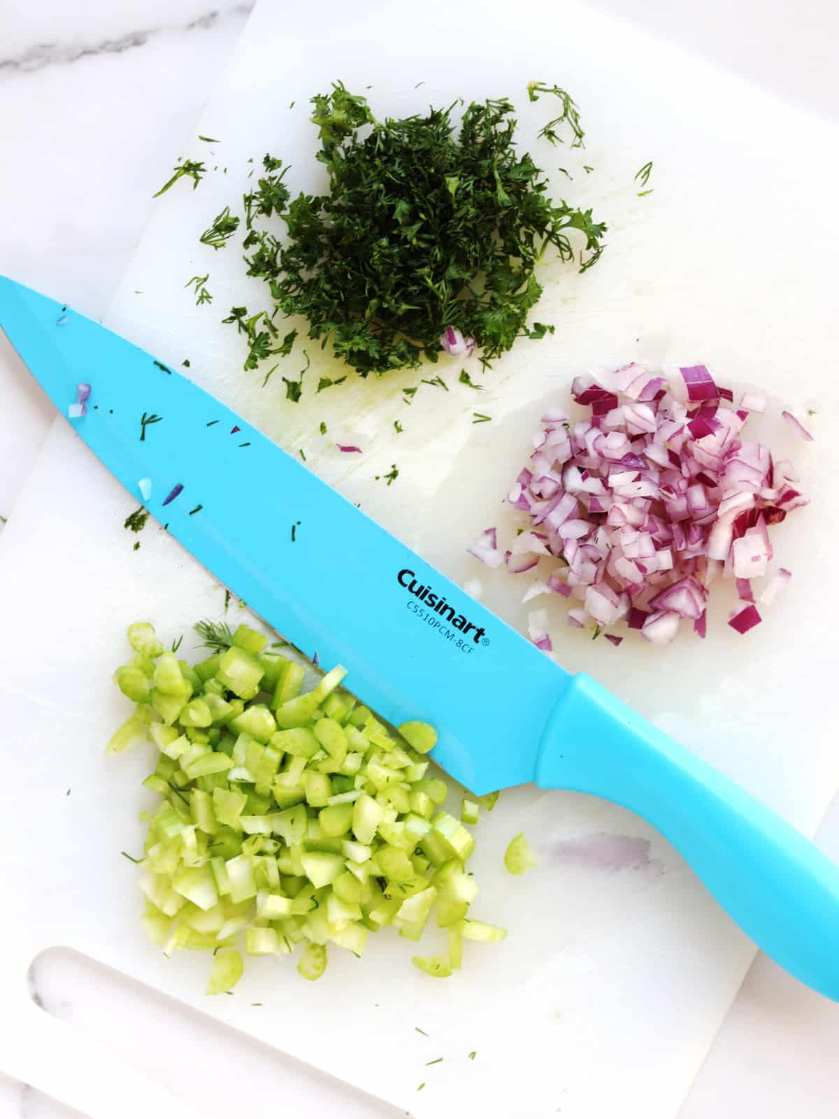 A blue-handled Cuisinart knife rests on a white cutting board with chopped fresh dill, diced red onion, and diced celery arranged in small piles.