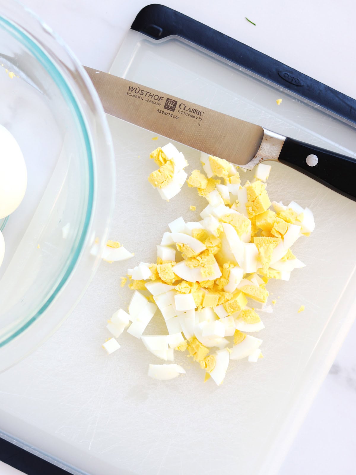 A knife rests on a cutting board beside chopped hard-boiled eggs. A glass bowl with more eggs is partially visible on the left side of the image.