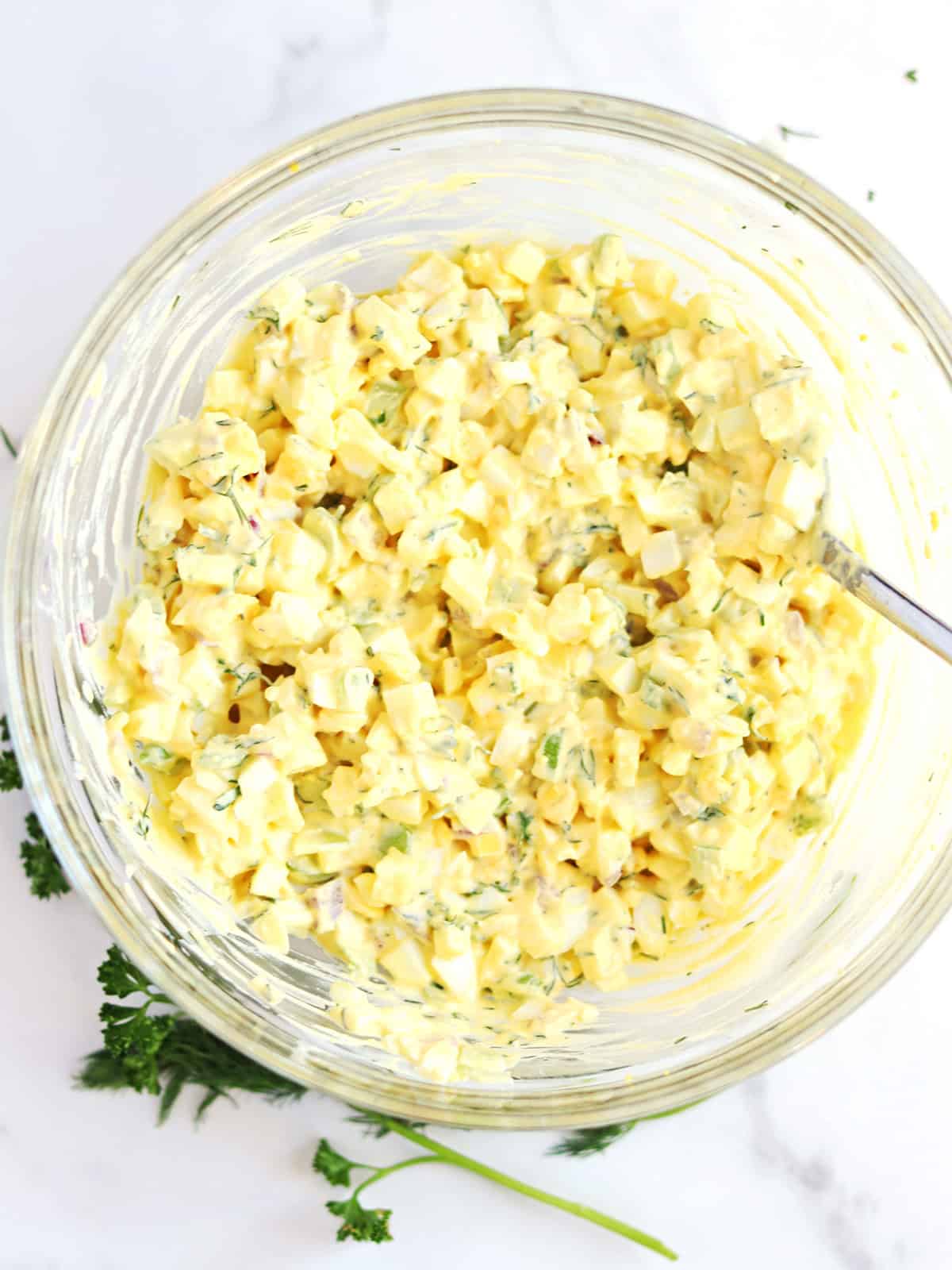 A glass bowl filled with creamy egg salad mixed with herbs, with a spoon inside. Fresh parsley is scattered next to the bowl on a white surface.