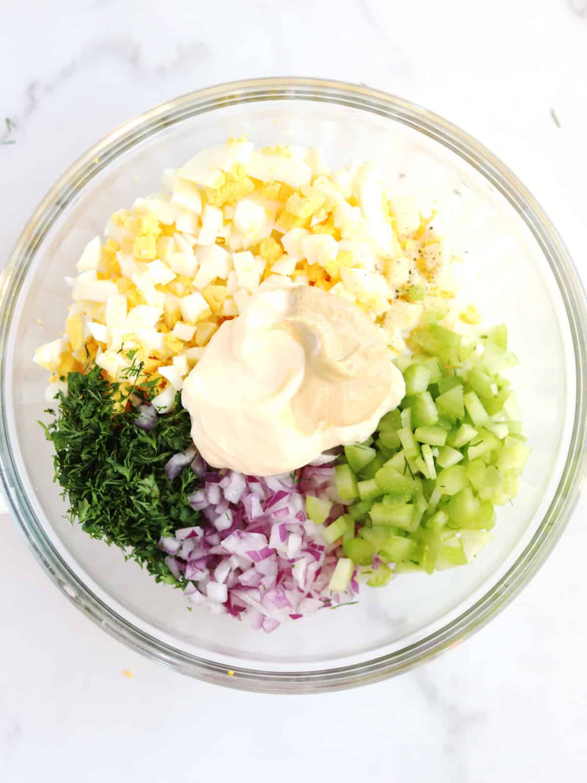 A glass bowl filled with chopped hard-boiled eggs, mayonnaise, mustard, chopped celery, diced red onion, and fresh dill, arranged in sections on a white marble surface.