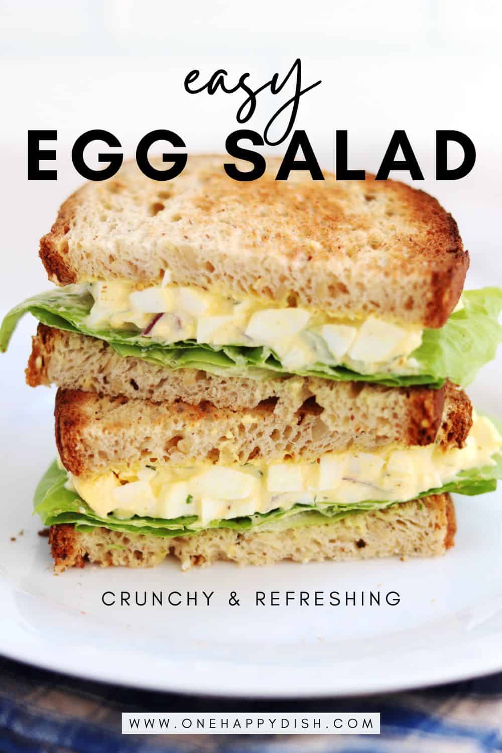A sandwich with egg salad and lettuce, made with toasted whole grain bread, is cut in half and stacked on a white plate, resting on a blue and brown cloth.
