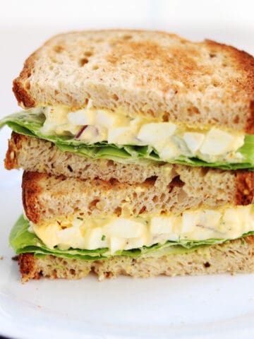 A plate with a stacked egg salad sandwich on whole wheat bread, featuring layers of chopped egg salad and green lettuce.