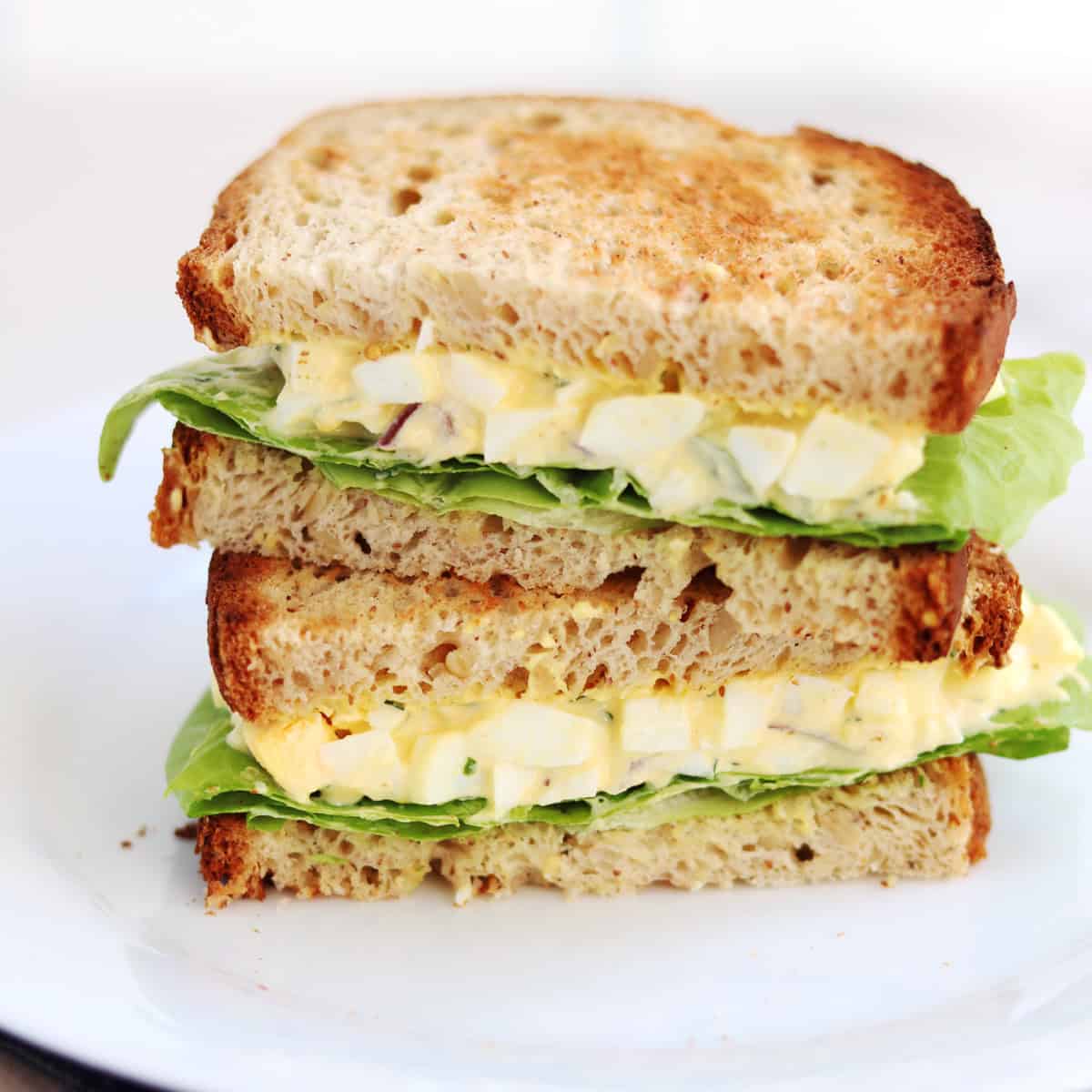 A plate with a stacked egg salad sandwich on whole wheat bread, featuring layers of chopped egg salad and green lettuce.