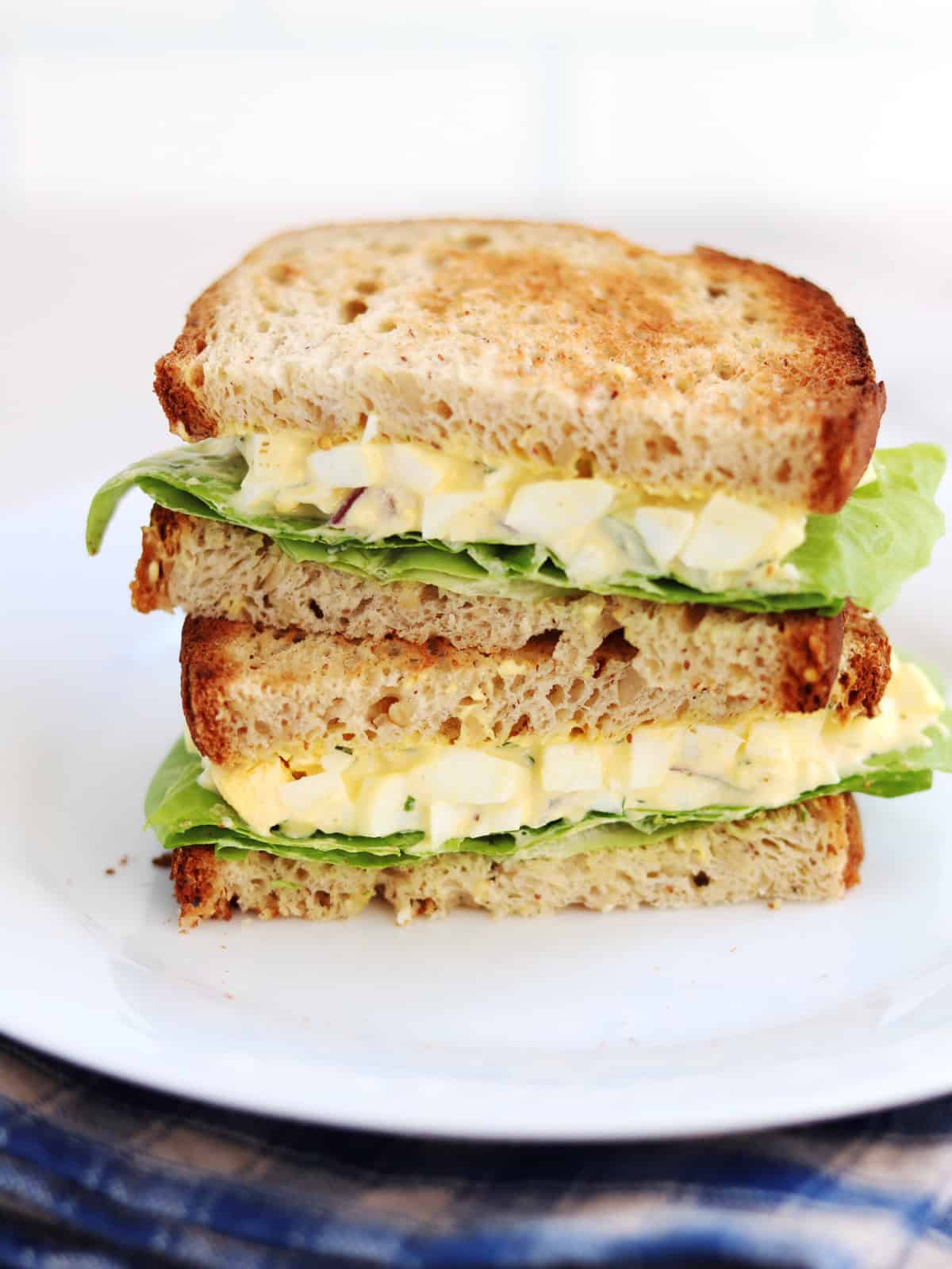 A stack of two egg salad sandwiches with lettuce on whole grain bread, placed on a white plate with a blue cloth underneath.
