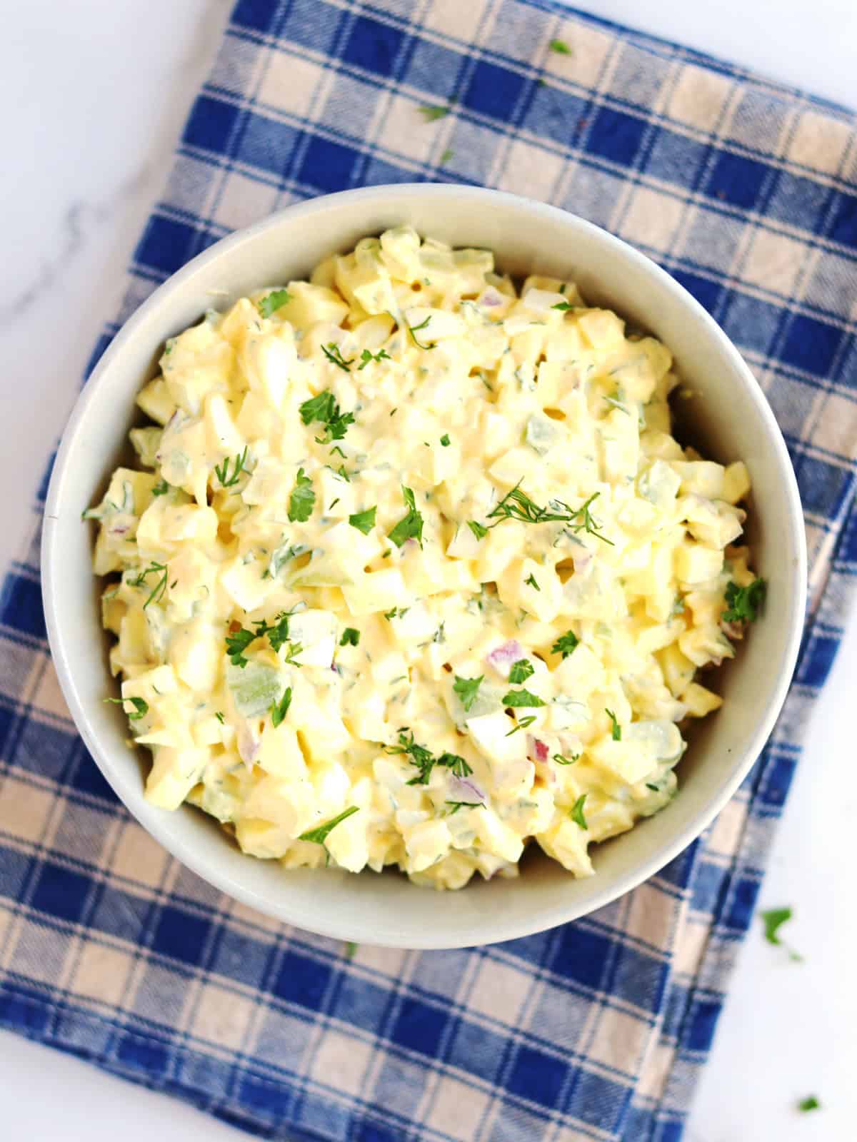 A bowl of creamy egg salad garnished with chopped herbs sits on a blue and beige plaid napkin on a white surface.