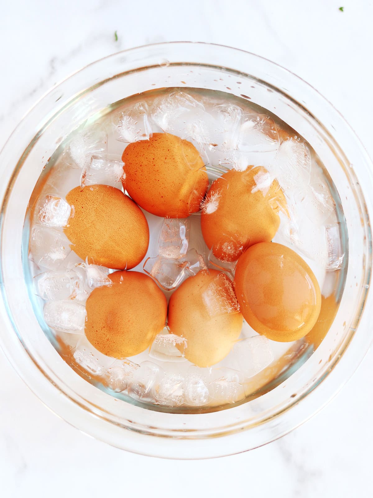 Five brown eggs are submerged in a glass bowl filled with ice cubes and water, seen from above on a white background.