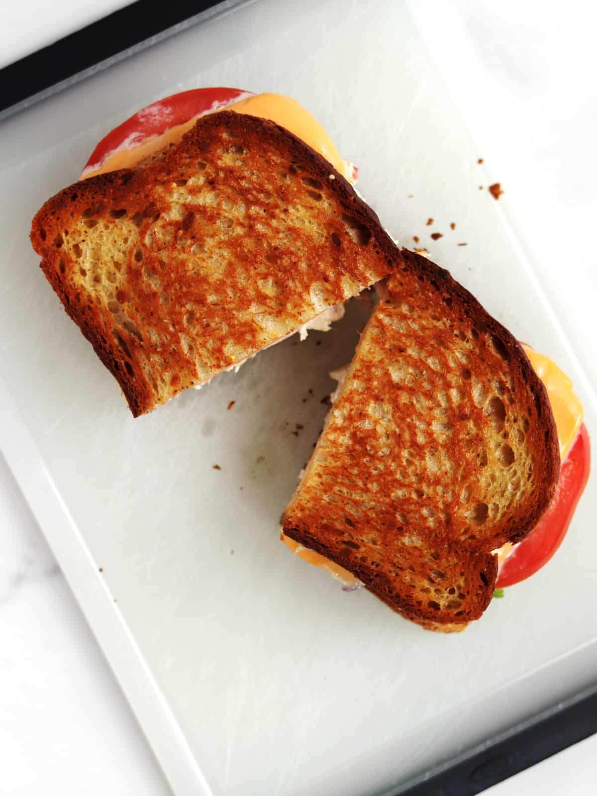 A grilled tuna melt sandwich cut in half on a white cutting board, showing golden-brown toasted bread and melted cheese and sliced tomatoes.