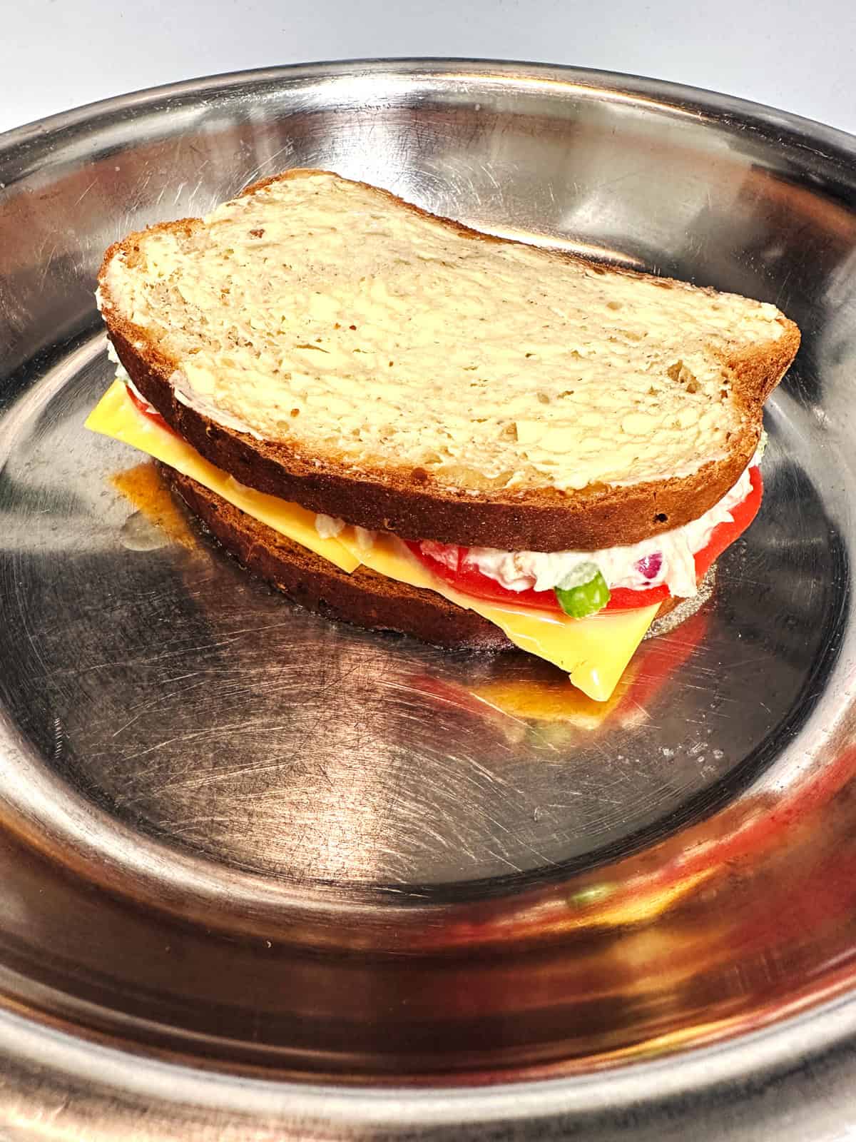 A prepared tuna melt sandwich in a stainless steel pan with cheese, tomato slices and tuna salad between slices of buttered bread.