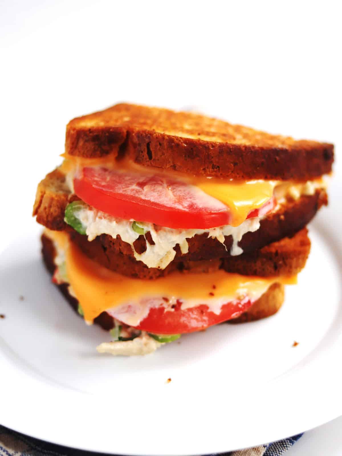 A grilled tuna melt sandwich with melted cheese, tomato slices and tuna salad between toasted bread, stacked on a white plate.