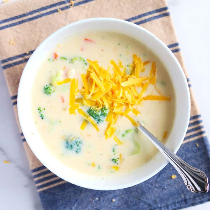 A bowl of creamy broccoli cheese soup topped with shredded cheddar cheese, with a spoon inside, placed on a striped beige and blue cloth.