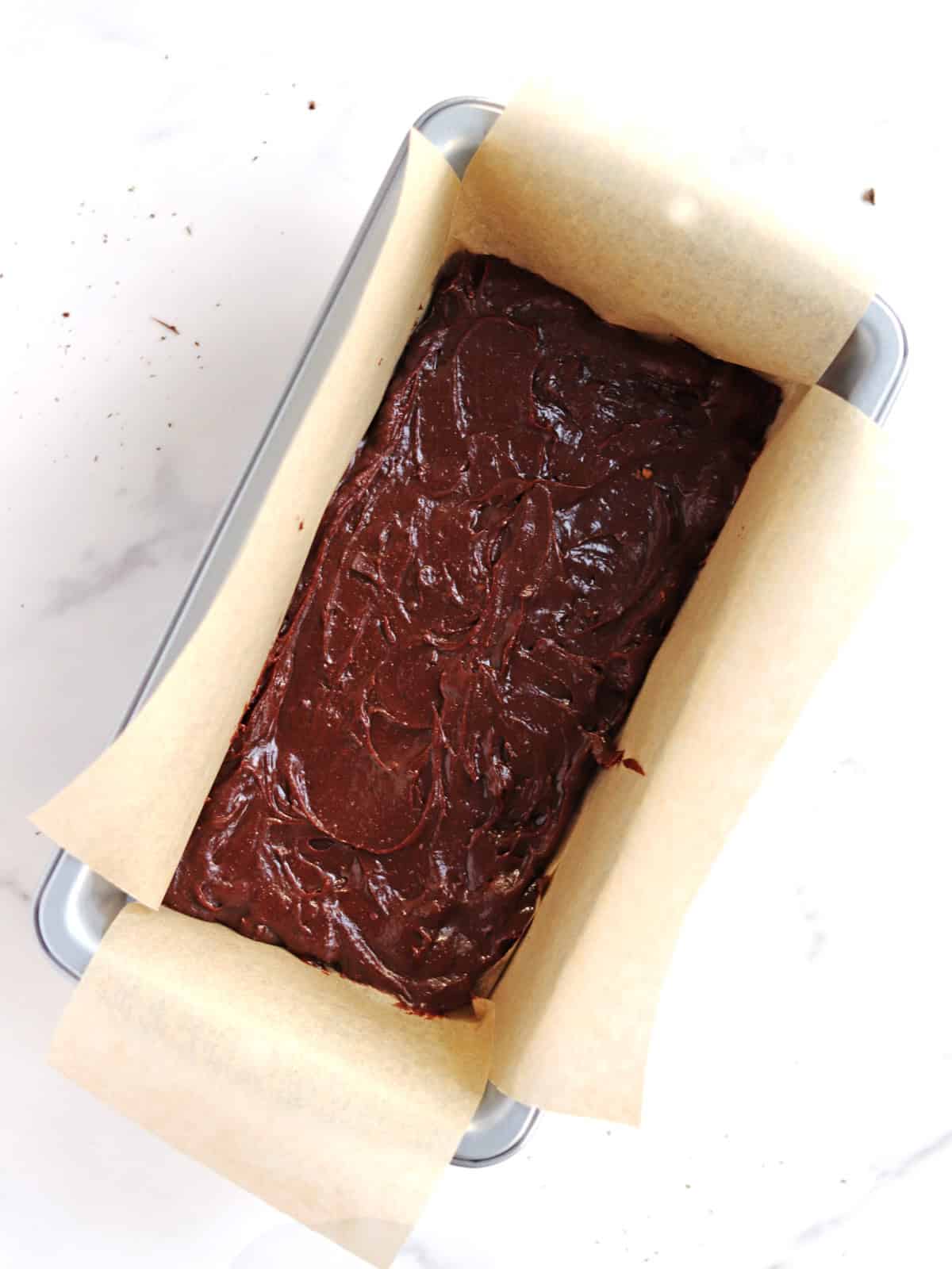 A loaf pan lined with parchment paper is filled with thick, swirled brownie batter, ready to be baked. The pan sits on a white surface.