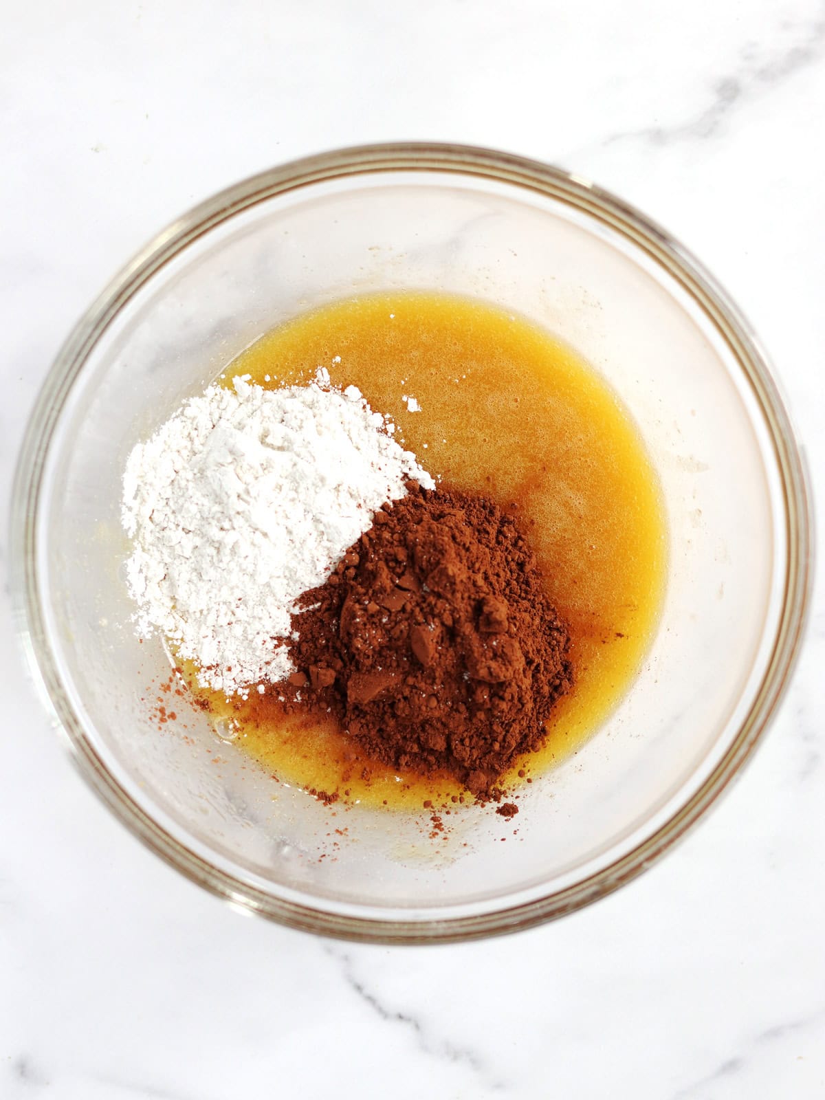A glass mixing bowl containing flour, cocoa powder, melted butter and sugar on a white marble surface, viewed from above.