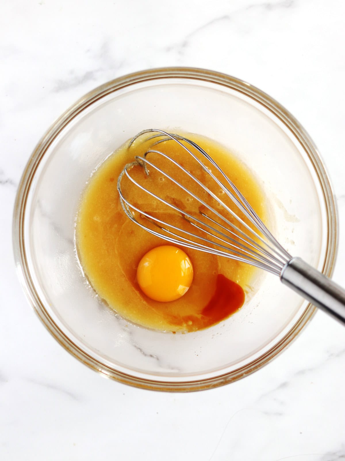 A glass bowl containing a raw egg, butter, sugar, and a bit of vanilla extract with a metal whisk resting inside, on a white marble surface.