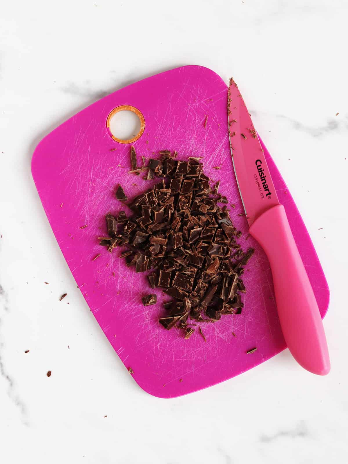 A pink cutting board with chopped dark chocolate pieces and a pink-handled knife on a white marble surface.