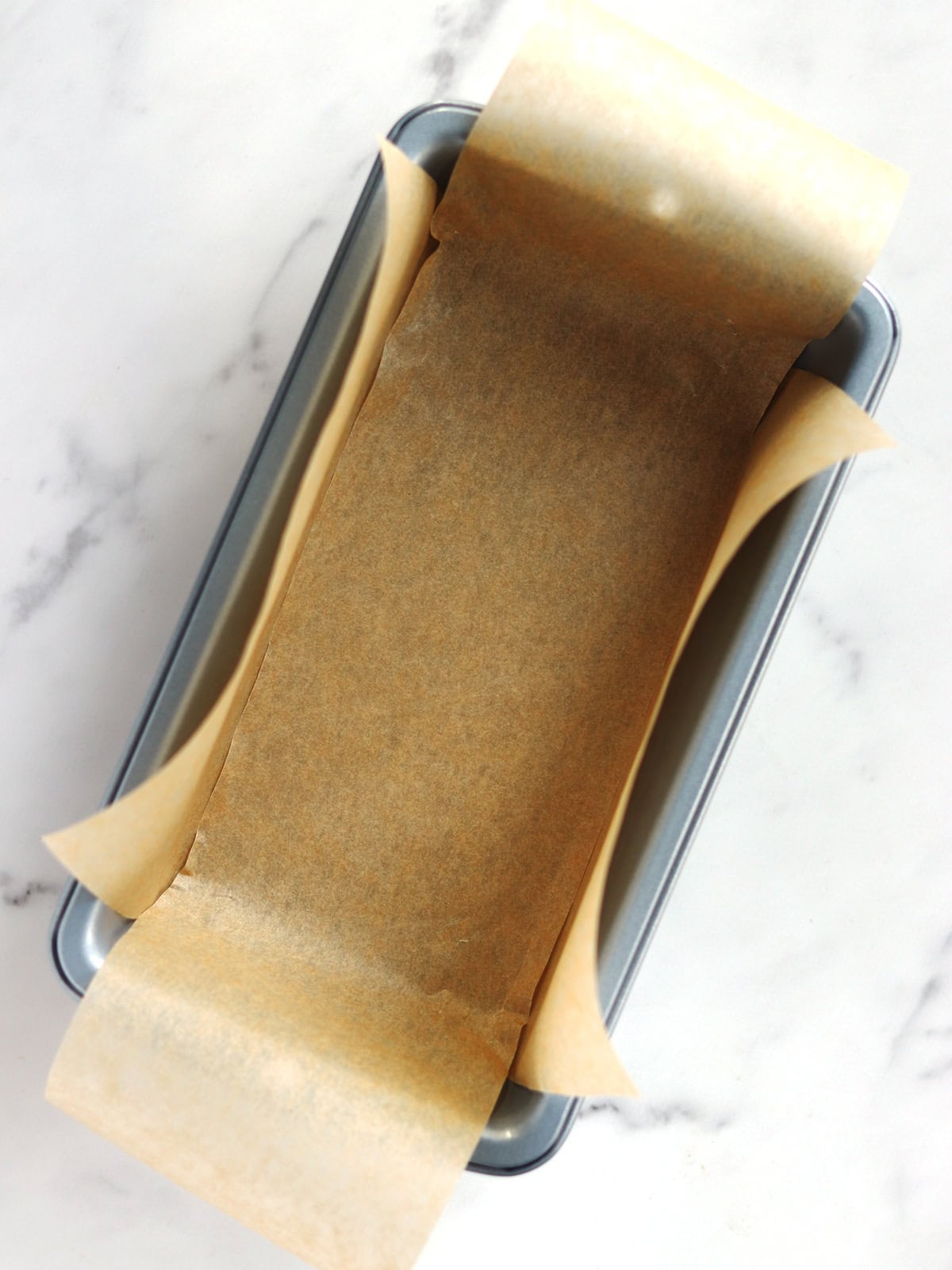 A metal loaf pan lined with parchment paper, placed on a white marble surface. The parchment paper extends beyond the edges of the pan for easy removal.