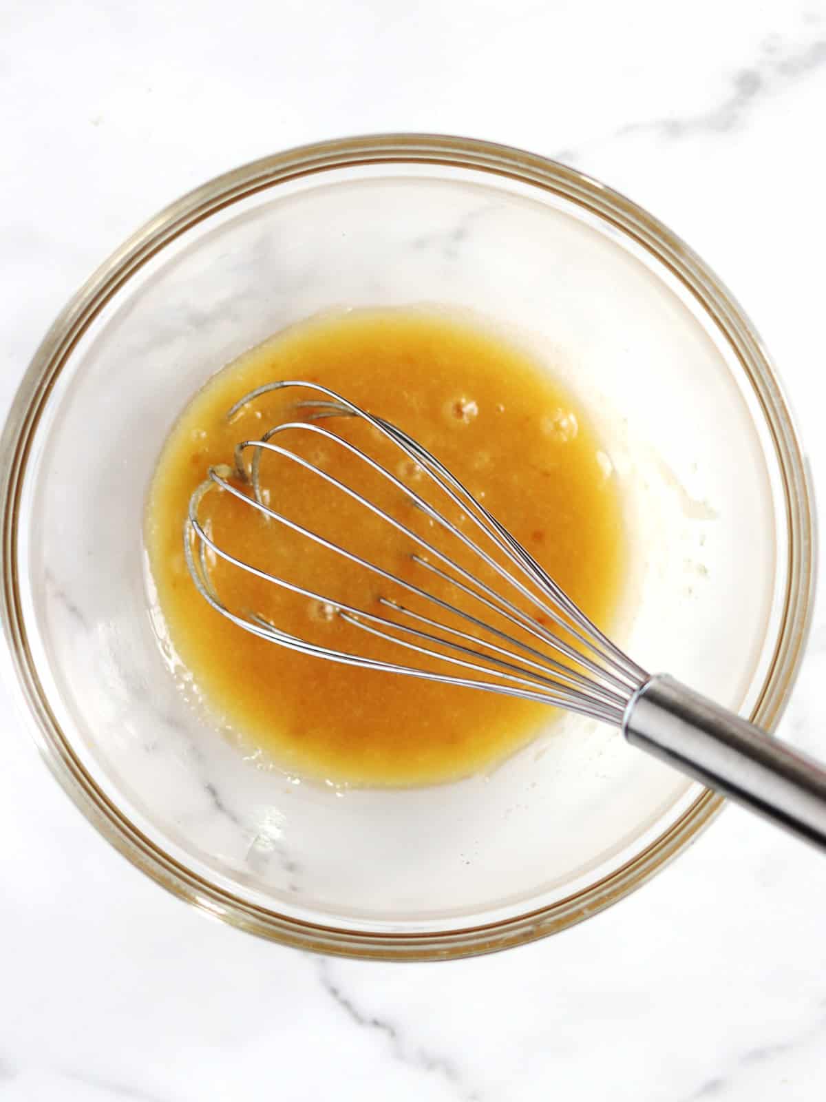 A metal whisk rests in a glass bowl containing a melted butter and sugar mixture on a white marble surface.