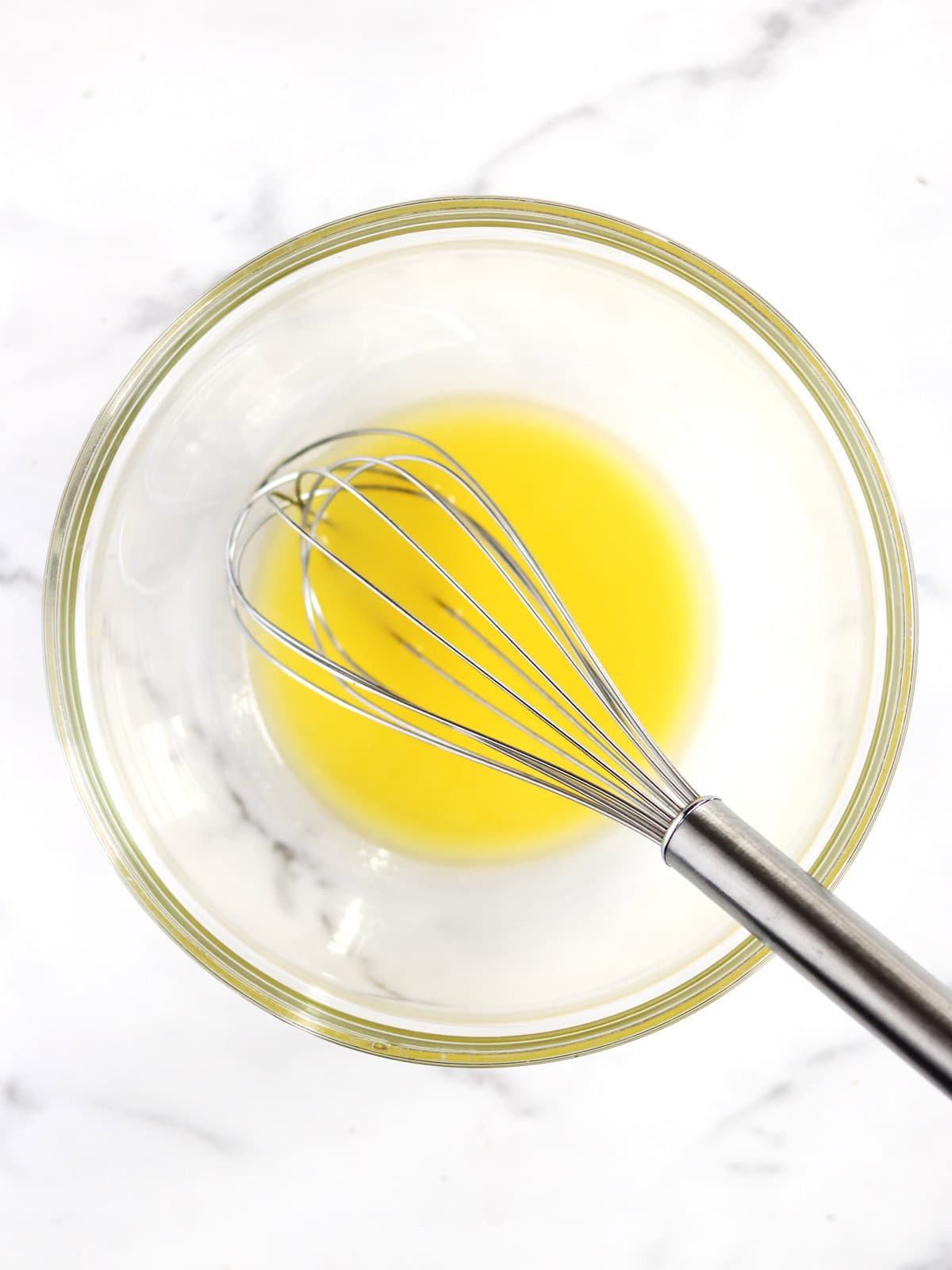 A metal whisk rests inside a clear glass bowl containing a small amount of melted yellow butter, set on a white marble surface.