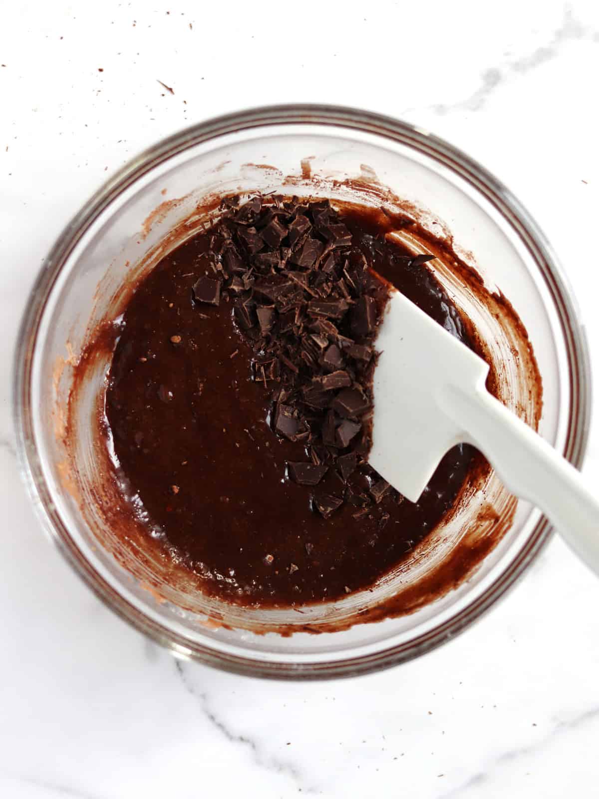 A glass bowl filled with chocolate brownie batter and chunks of chopped chocolate, being mixed with a white spatula on a white marble surface.