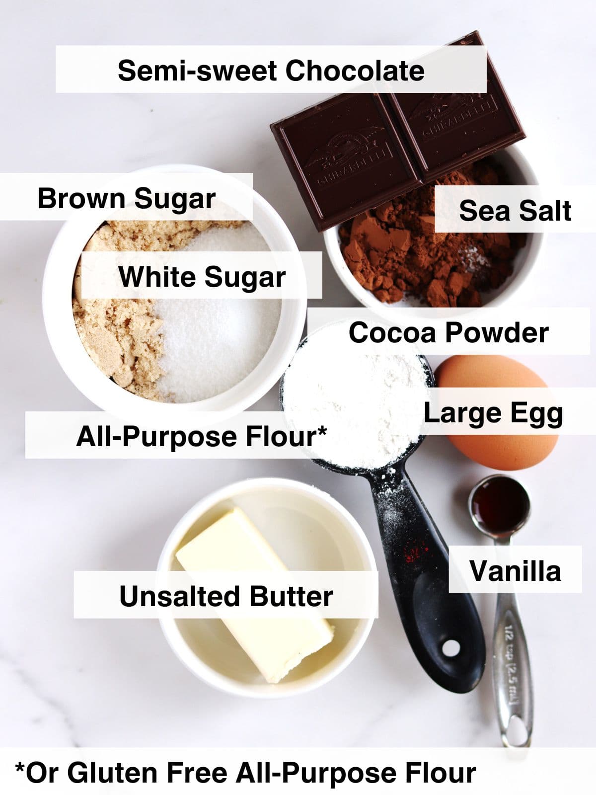 A flat lay of labeled baking ingredients: semi-sweet chocolate, brown sugar, white sugar, sea salt, cocoa powder, all-purpose flour, a large egg, vanilla, and unsalted butter on a white surface.