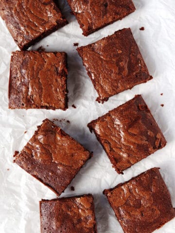 Eight square chocolate brownies are arranged in a scattered pattern on crinkled white tissue paper.