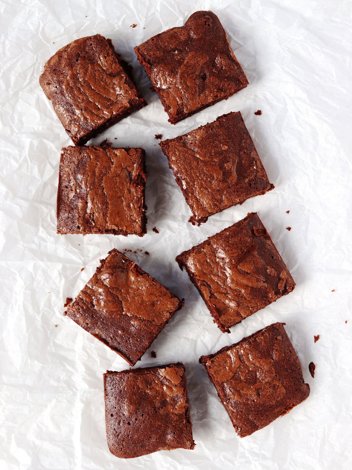 Eight square chocolate brownies are arranged in a scattered pattern on crinkled white tissue paper. The brownies have a rich, glossy top and fudgy texture.
