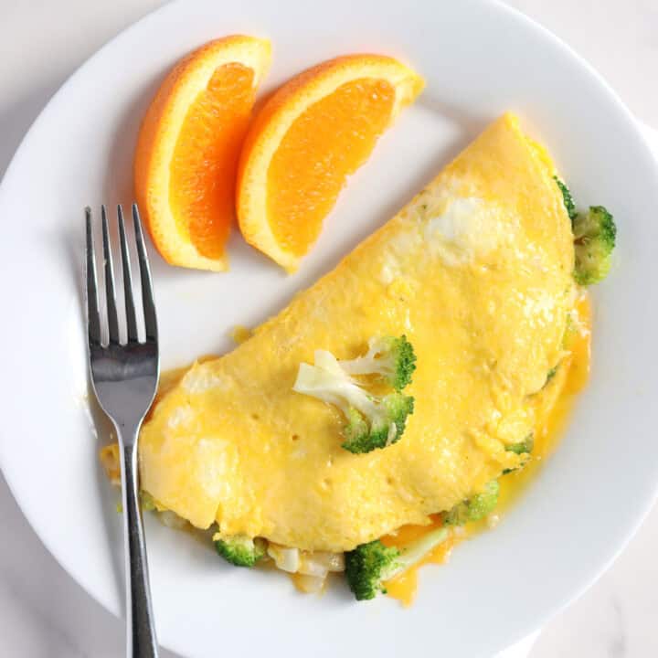 A broccoli and cheese omelette on a white plate, garnished with a broccoli floret. Two orange slices and a fork are placed beside the omelette.