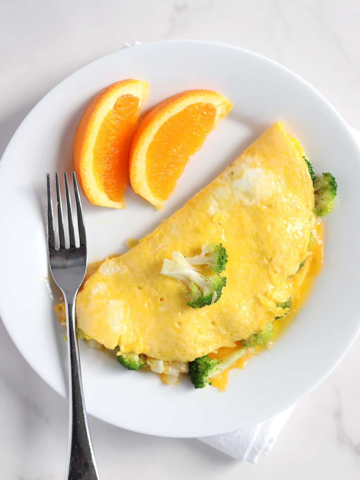 A plate with a cheese and broccoli omelette, garnished with a small broccoli floret, two orange slices, and a fork resting on the side. The setting is bright with a white background.