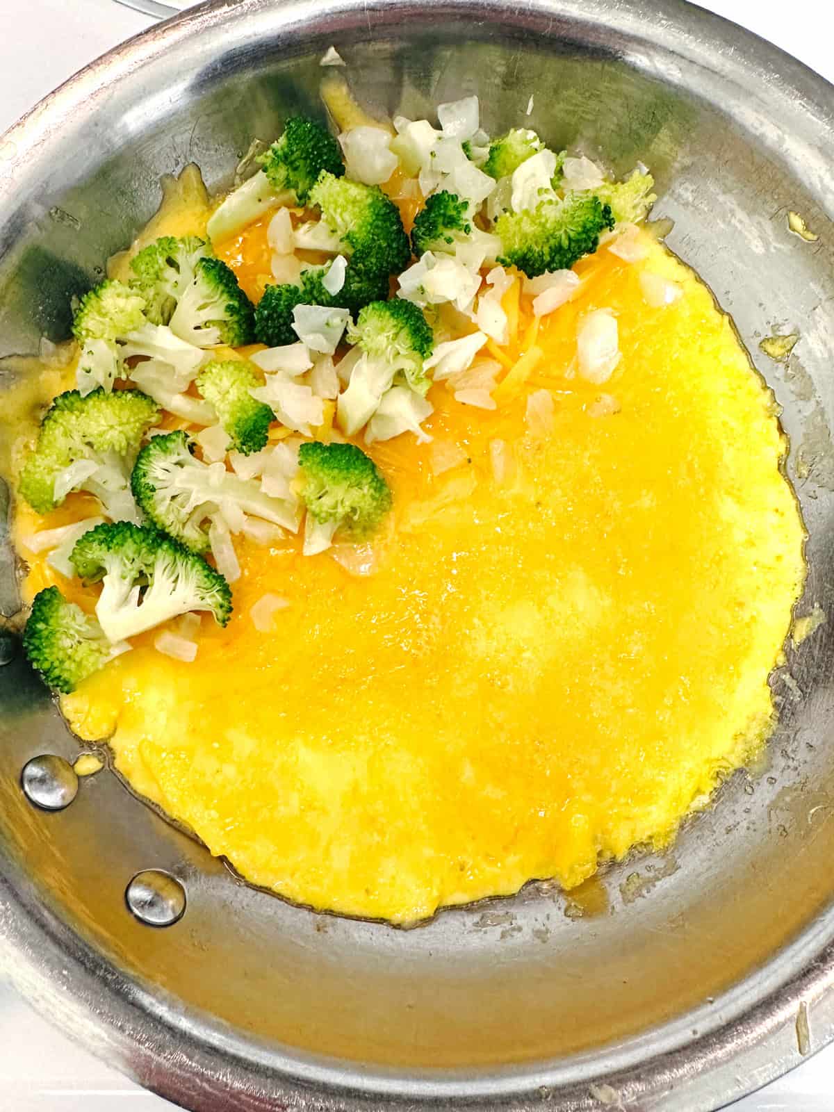 A partially cooked omelet in a skillet, topped on one side with chopped broccoli and diced onions. The eggs are still soft and yellow, while the vegetables are fresh and lightly cooked.