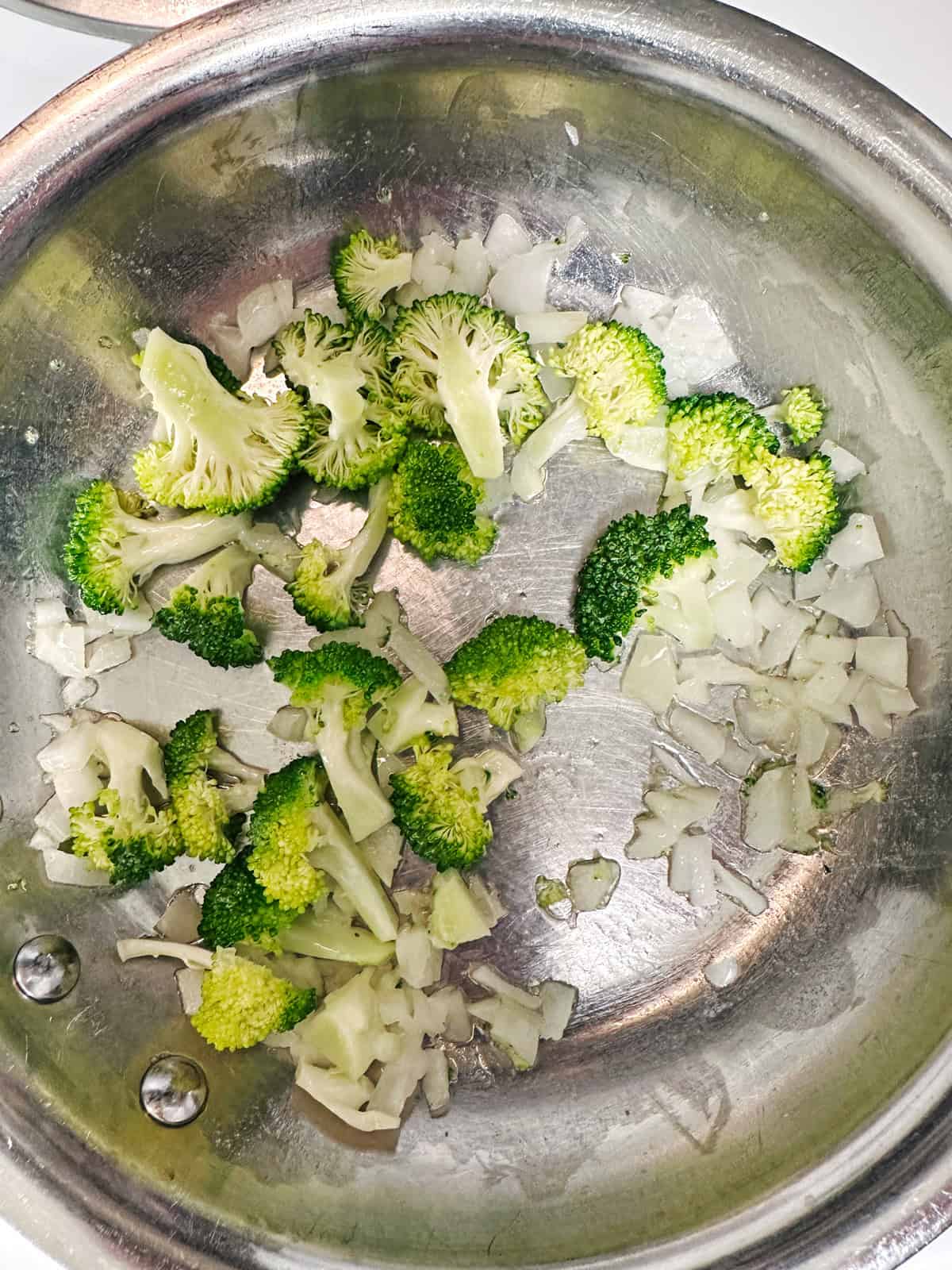 Chopped broccoli florets and diced onions cooking in a stainless steel pan.