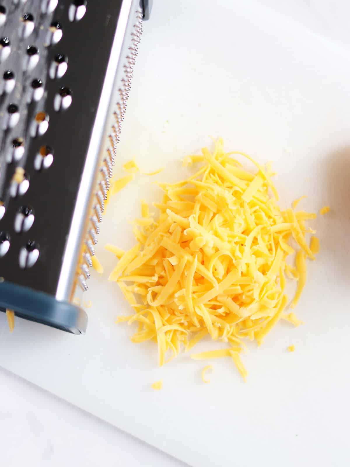 A pile of freshly grated cheddar cheese sits on a white cutting board next to a metal box grater.